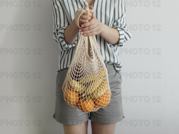 Mesh bag with fruits in female hands. Stylish young woman hold mesh shopping bag on light gray wall
