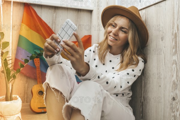 Young millennial hippie woman sitting on balcony using mobile phone. LGBTQ rainbow flag on