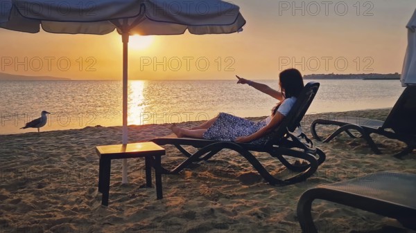 Young woman sitting on a sunbed at the beach pointing forefinger to the sunrise over the horizon.