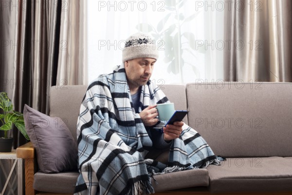 Middle age male model in hat and plaid hold coffee cup sitting on the sofa while using his