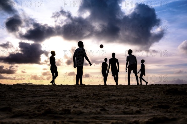 Group of people enjoying a football match on the beach at sunset, silhouettes against a backdrop of