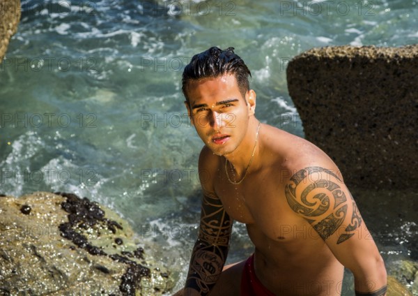 Attractive young shirtless athletic man standing in water by sea or ocean shore, wearing shorts,