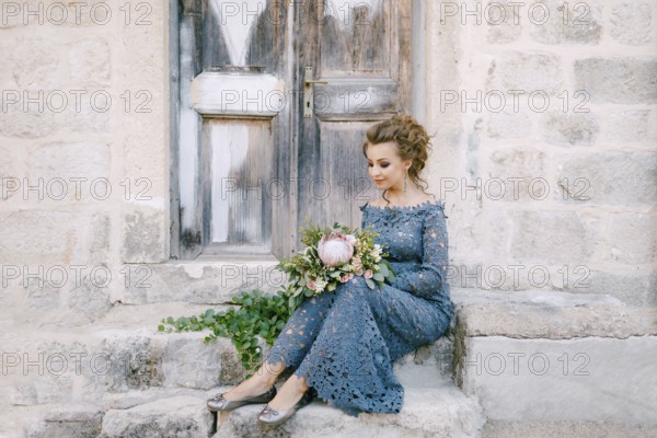 A bride in a stylish blue dress with a bouquet in her hands sits on the steps near an old wooden