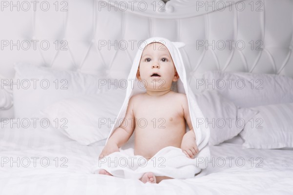 Adorable newborn baby boy with brown eyes sitting wrapped in a white towel with ears in light bed.