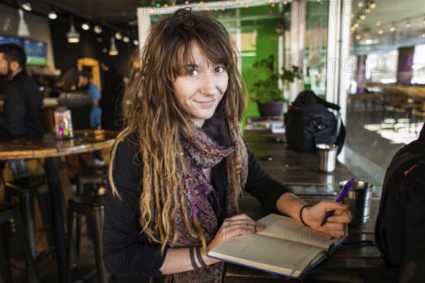 Portrait of smiling beautiful girl with dreadlocks and tattoos. Writing on a notebook with a pen in