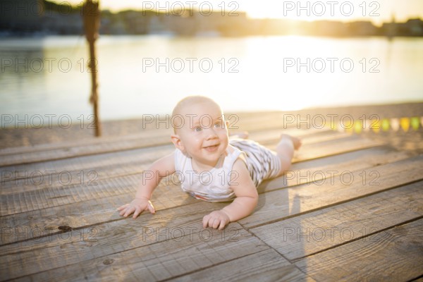 A child a boy, one year old, blond man lies on his stomach on wooden dock, pier in striped clothes,