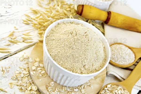 Oat flour in white bowl, oatmeal and bran in wooden spoon, stalks of oats, a rolling pin and a