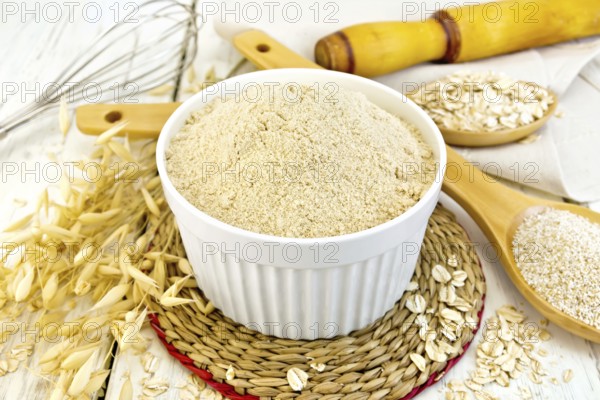 Oat flour in white bowl, oatmeal and bran in wooden spoon, stalks of oats on the background of