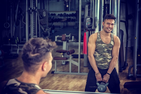 Attractive young athletic man exercising in gym, working out using kettlebell, looking at camera