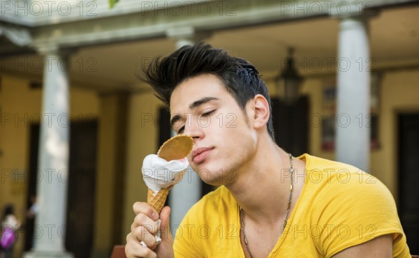 Funny attractive young man sitting, wearing yellow T-shirt while eating a tasty vanilla ice cream