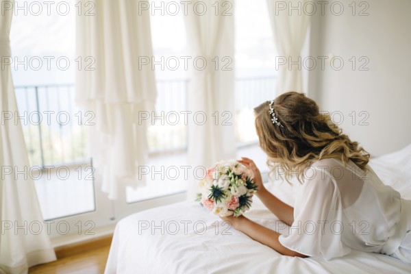 A bride in a gentle peignoir with a bouquet in her hands laying on the bed by the window with white
