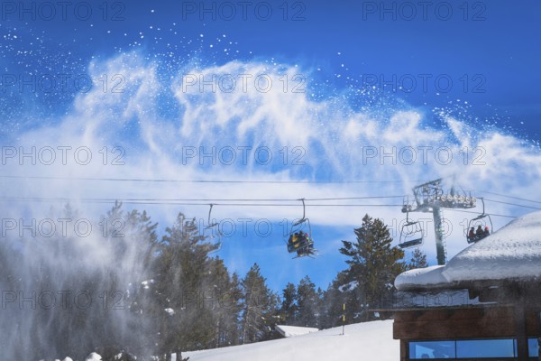 Snow cannon spreading fresh snow powder on a ski slope. Skiers riding up on chair lift. Winter ski