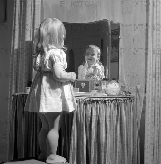 In the 1950s. A little girl is standing in front of her mothers makeup table and mirror and puts on