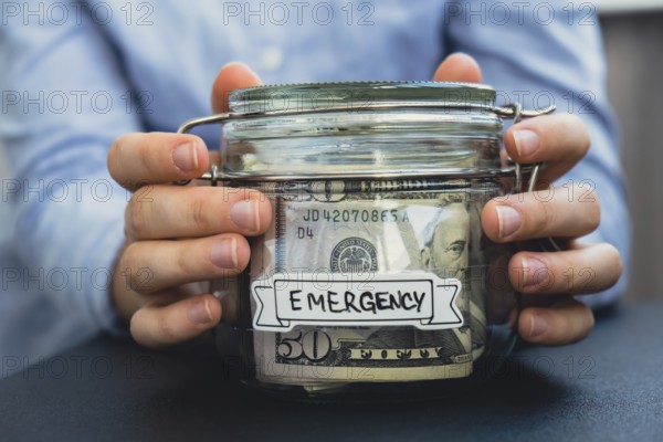 Female hands Saving Money In Glass Jar filled with Dollars banknotes. EMERGENCY transcription in