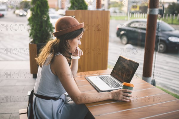 Beautiful young caucasian woman sitting on a terrace in a cafe in summer at a wooden table in a hat
