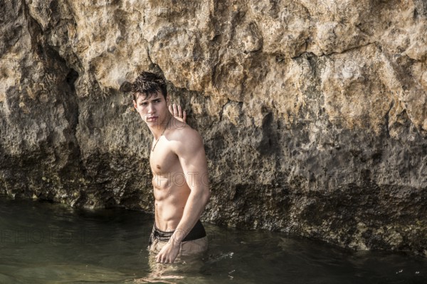 Attractive young shirtless athletic man standing in water by sea or ocean shore, wearing shorts,