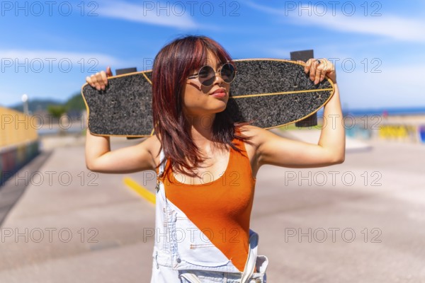 Portrait of a urban confident asian woman holding a skateboard on shoulders standing in an urban
