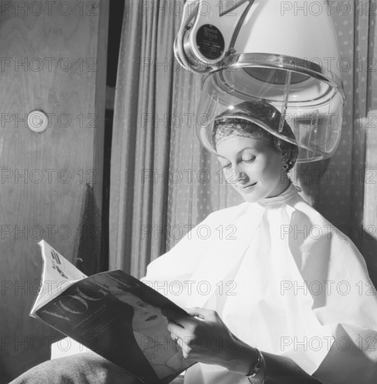 Hair dryer in the 1950s. A woman is sitting comfortably and reading Vogue magazine, while drying