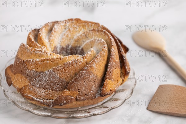 Fresh marble bundt cake, cake with icing sugar next to a rustic wooden spoon on a light-coloured