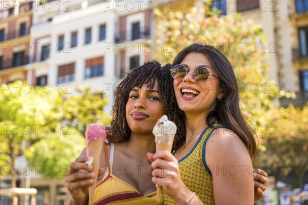 Two smiling girls are eating ice cream cones, hugging each other in a city center during a sunny
