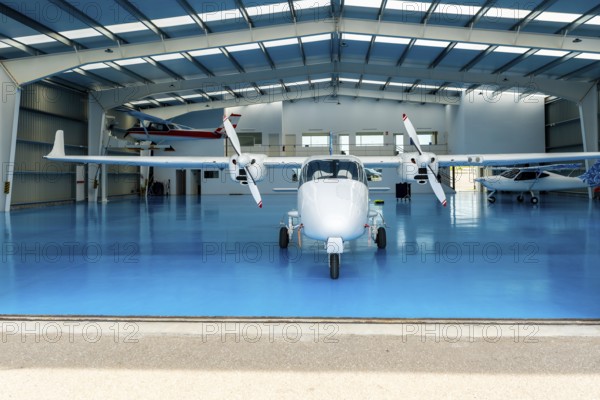 White twin engine airplane parked inside a spacious hangar, featuring a bright blue floor and