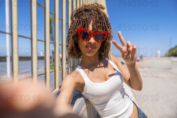 Young woman with heart shaped sunglasses takes a selfie while making the peace sign, leaning