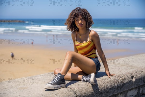 Stylish young woman wearing casual summer clothes sits on a beach wall, enjoying the ocean view