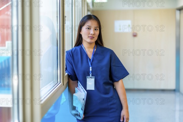 Confident young medic holding a clipboard and wearing blue scrubs is standing next to a window in a