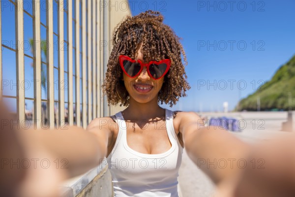 Smiling young woman with curly hair taking a selfie with heart shaped sunglasses on a sunny beach