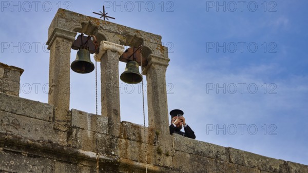 Priest next to bells on a wall in front of a blue sky, Monastery of St John the Theologian,