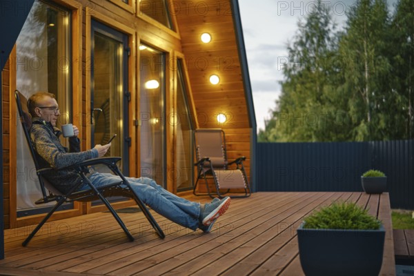 Middle-aged man sits with a cup of tea in the evening on the terrace illuminated by outdoor wall