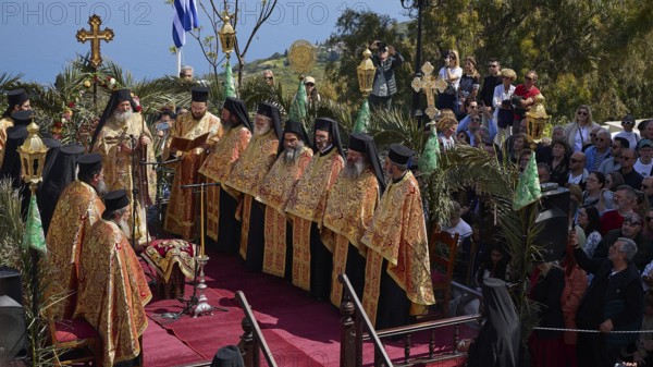 Priests in golden robes lead a religious ceremony on a decorated stage in front of a Greek flag and