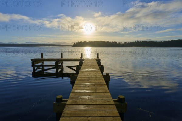 A jetty leads into a lake, the sun shines brightly and is reflected in the water, Lake Mahinapua,