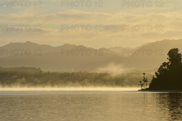 A calm lake at dawn with mist, surrounded by silhouetted mountains, Lake Mahinapua, Ruatapu, South
