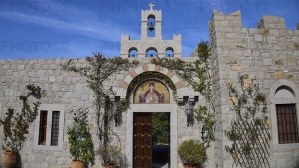 Stone building with bell tower and religious mosaic, surrounded by climbing plants, woman's