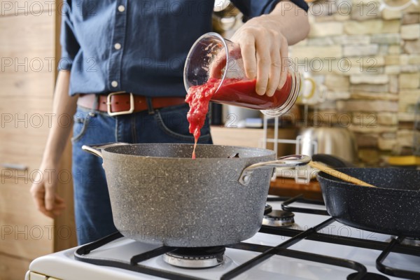 A person pours tomato sauce from a jar into a pot on the stove. The kitchen features modern decor