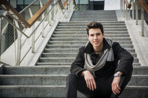 Trendy handsome young man in winter fashion sitting on a long staircase leading to an urban