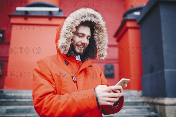 Handsome young male student with toothy smile and beard stands on red wall background, facade of