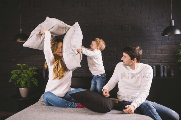 Parents with smiling child playing pillow fight on bed, happy family having fun in bedroom,