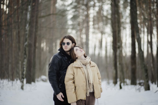 Couple in a snowy forest with the woman leaning on the man, both in winter attire among towering