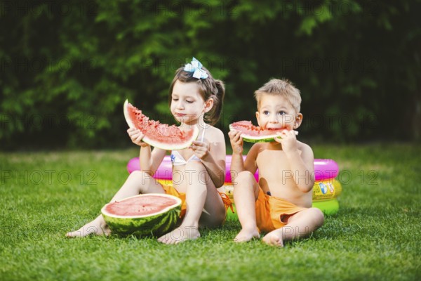 Funny little kids brother and sister eating watermelon on green graß near inflatable pool in yard