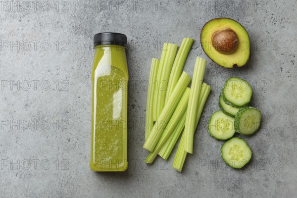 Bottle of green vegetable smoothie with fresh celery sticks, cut avocado, cucumber on gray concrete