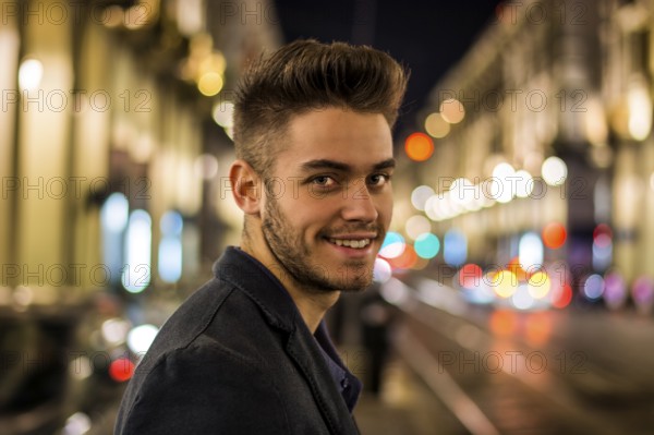 Attractive young man portrait at night with city lights behind him, smiling and looking at camera