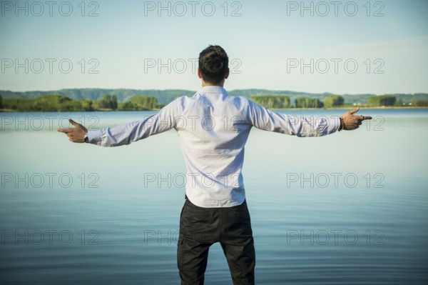 Young man in shirt, outdoor with arms spread open enjoying freedom in front of lake, seen from the