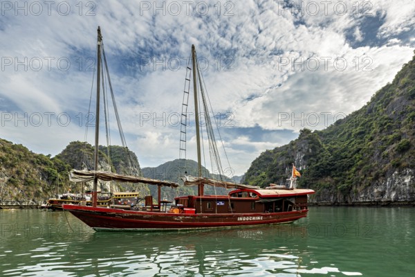 Traditional wooden boat sails on turquoise water between green hills under a cloudy sky, sailing