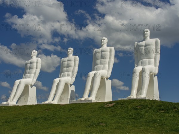 Four large white sculptures of seated men on a green meadow under a blue sky with clouds, esbjerg,