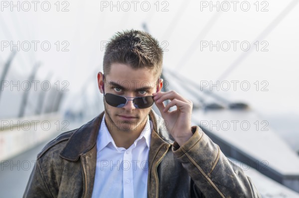 Handsome trendy young man standing on a sidewalk, wearing sunglasses and a fashionable leather
