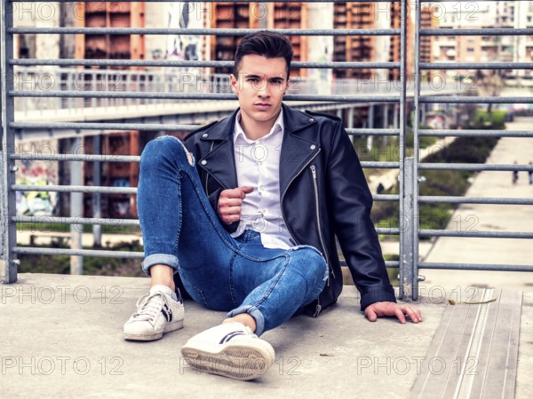 One handsome young man in urban setting in European city, sitting, wearing black leather jacket and