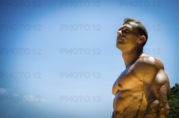 Handsome young muscle man at the seaside, outdoors, showing muscular pecs and torso in front of the