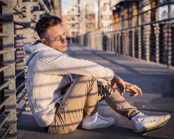 Handsome trendy young man, sitting in city setting at sunset wearing a fashionable sweater or
